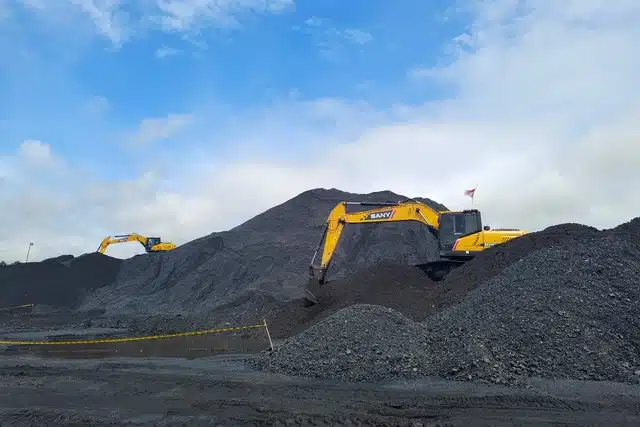 Dua ekskavator kuning beroperasi di tumpukan batu bara pada area stockpile tambang dengan langit cerah berawan